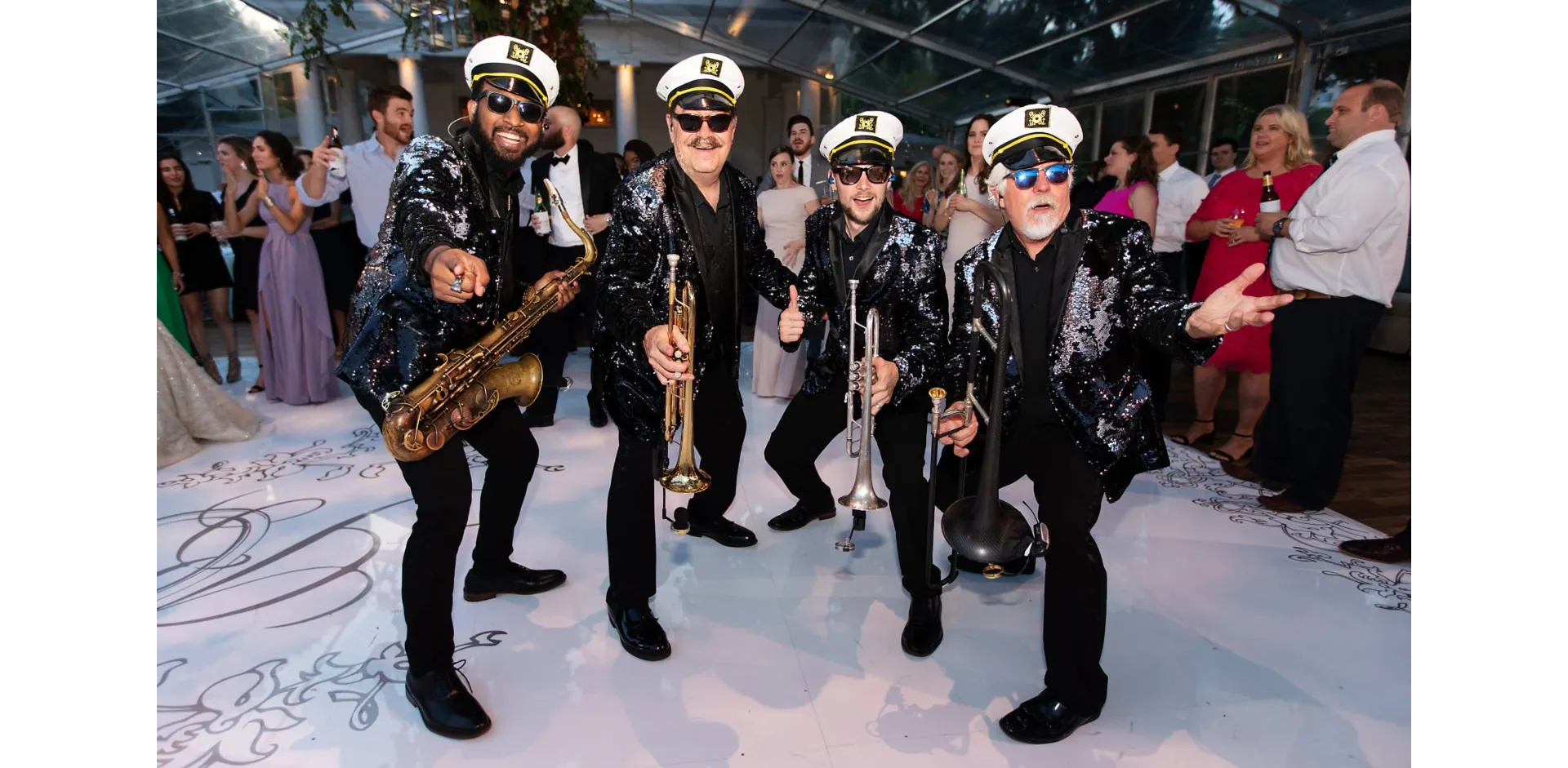 Four musicians in matching sequin jackets and captain hats pose with instruments at a lively event.