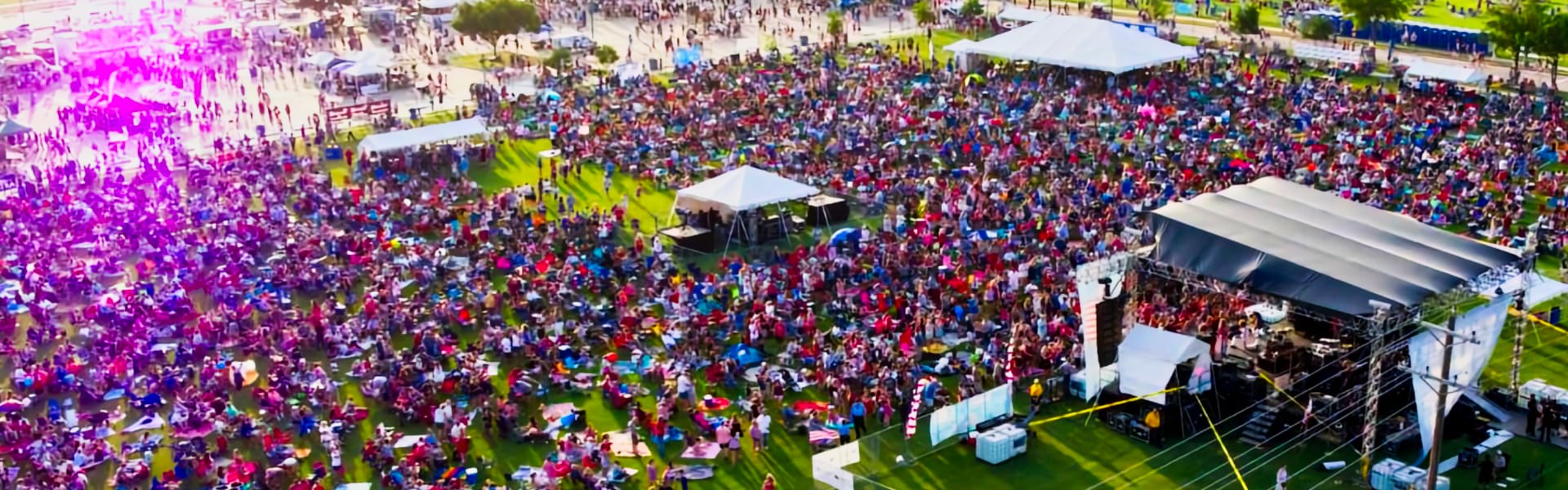 Large crowd gathered on grass in front of an outdoor concert stage on a sunny day.