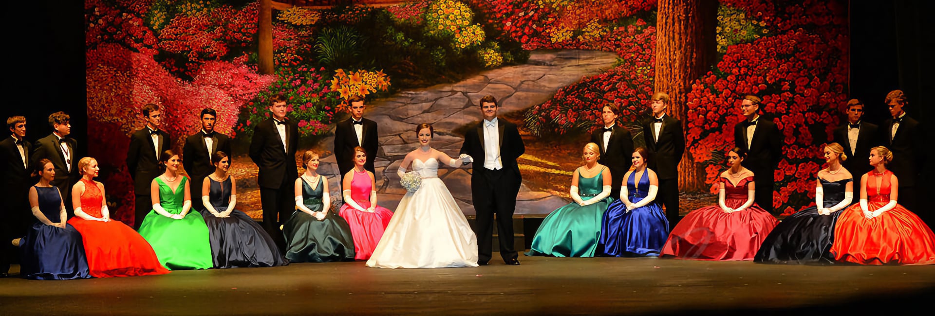 People in formal attire on stage, women in colorful gowns, men in tuxedos, autumn backdrop.