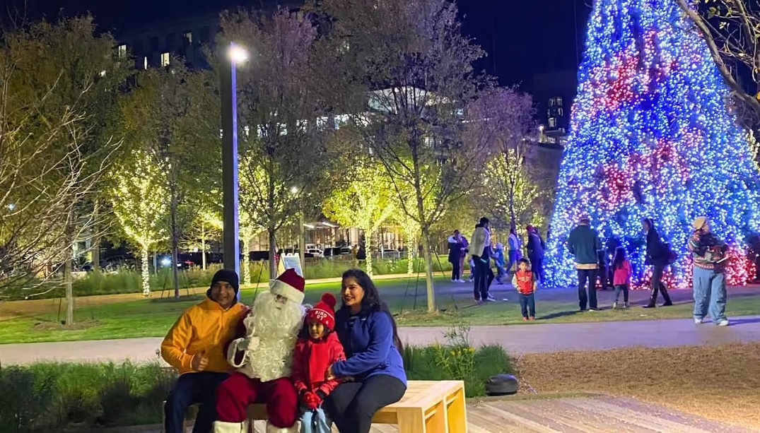 Family sitting with Santa near a brightly lit Christmas tree in a park at night.