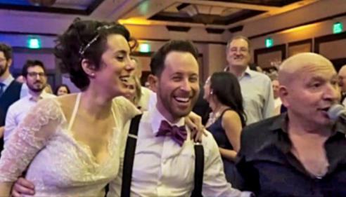 Bride and groom laughing with a singer at a lively wedding reception.