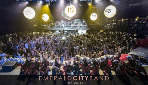 Large crowd with a band posing on stage under bright concert lights.