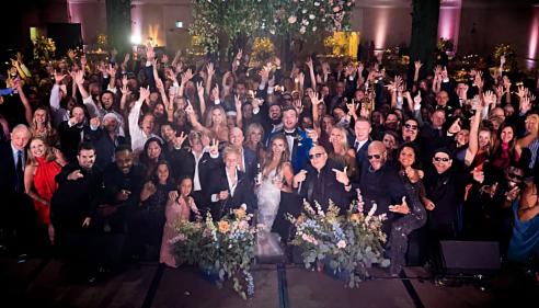 Large group of people celebrating at a wedding reception, raising hands and smiling.