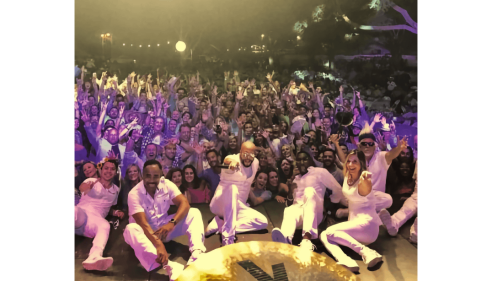 Performers in white pose on stage in front of a cheering crowd under purple lights.