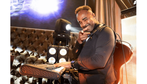 DJ smiling and pointing while playing music at a club with stage lights behind him.