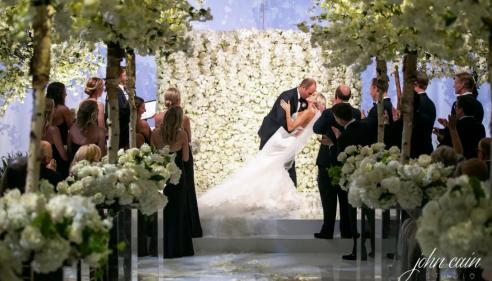 Bride and groom kiss at flower-filled wedding ceremony, surrounded by applauding guests.