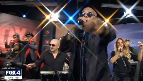 Band performing live on TV set, lead singer reaching forward, bright stage lights shine.