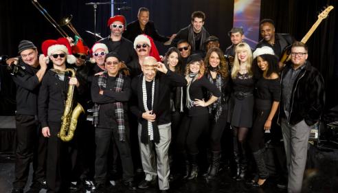 Band members in black outfits and Santa hats pose together on stage.