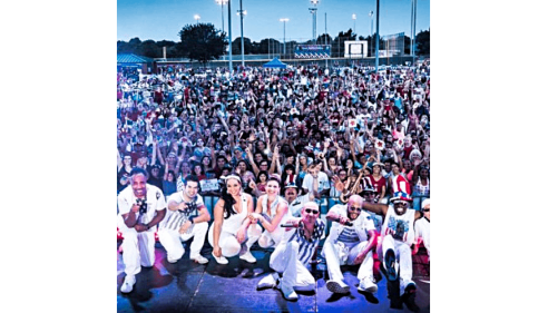 Performers in white outfits pose on stage with a large, cheering crowd behind them outdoors.