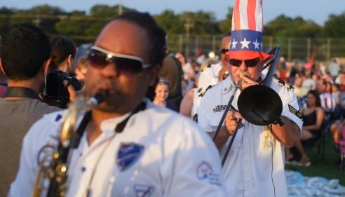 Musicians play brass instruments outdoors, one wearing a tall American flag hat.