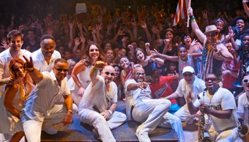 Musicians in white posing on stage with an enthusiastic crowd in the background.