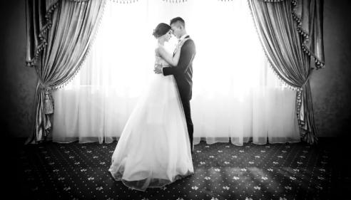 Bride and groom embrace in front of a bright window with elegant curtains.
