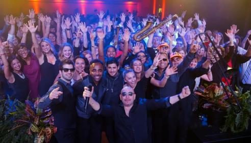 Large group of people at a lively party, smiling and raising their hands.