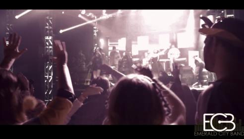 Concert crowd raising hands under bright stage lights, people silhouetted in the foreground.