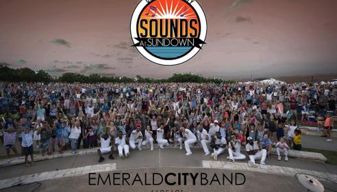 Large outdoor concert crowd with performers in white on stage at sunset.
