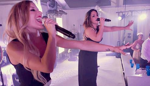 Two women in black dresses singing passionately on stage with microphones.