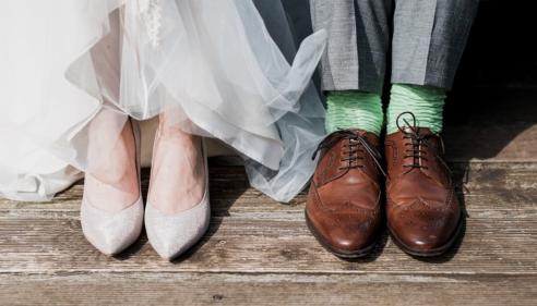 Bride’s white shoes beside groom’s brown shoes and green socks on wooden floor.