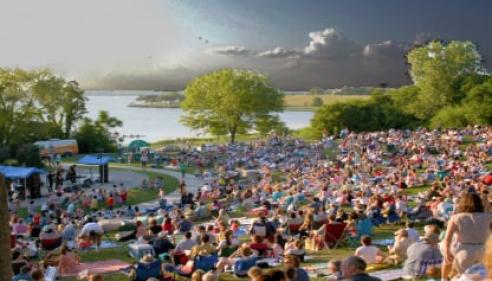 Large crowd sitting on grass at an outdoor concert near a lake under a partly cloudy sky.