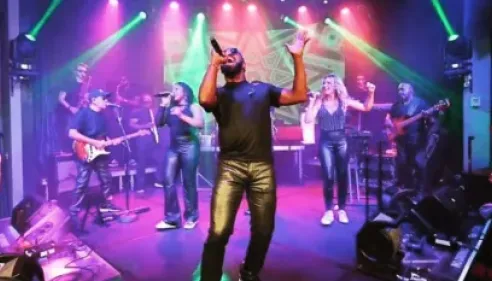 Band performing energetically on stage with colorful lights and singers dancing.
