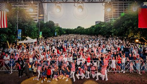 Large crowd at an outdoor event, many people wearing red, white, and blue, flags displayed.