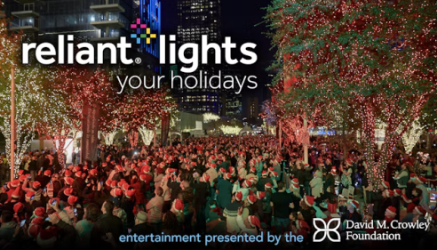 Large crowd in festive hats at a holiday lights event, trees lit with colorful lights.