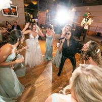 Bridesmaids in green dresses and a bride dance with a lively singer at a wedding reception.