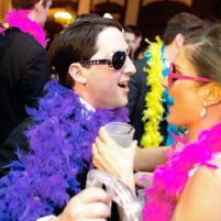 People in colorful feather boas and sunglasses dancing at a lively party.