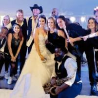 Bride and groom posing with friends in formal attire, smiling under festive lights.