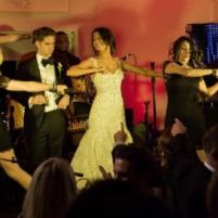 Bride, groom, and bridesmaids dancing joyfully on stage at a wedding reception.