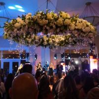 Crowd of people dancing under a large floral chandelier with blue and purple lighting.