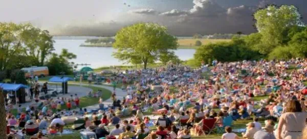 Large crowd gathered on a grassy hill near a lake, enjoying an outdoor event.