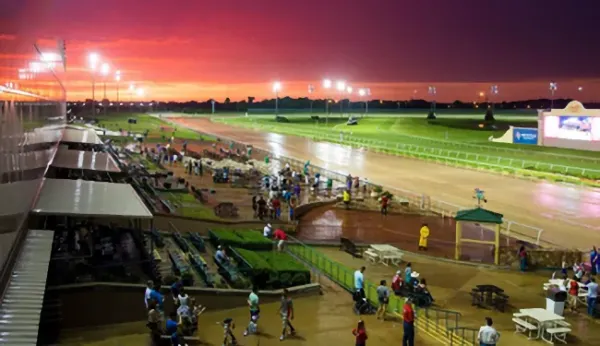 Crowd gathered by a racetrack at sunset under stadium lights.