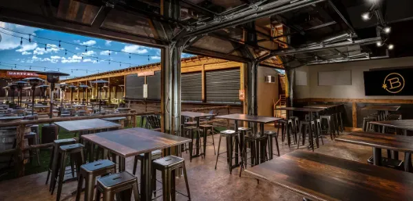 Modern indoor-outdoor bar with wooden tables, stools, and string lights overhead.
