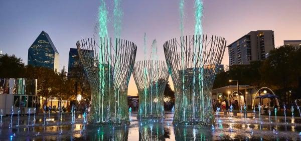 Colorful water fountains lit up at dusk in a city plaza with buildings in the background.