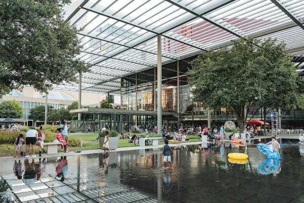 Children and families playing near a shallow reflecting pool in an outdoor urban plaza.