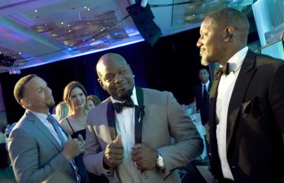 Three men in suits and a woman in a formal dress talking at a gala event.