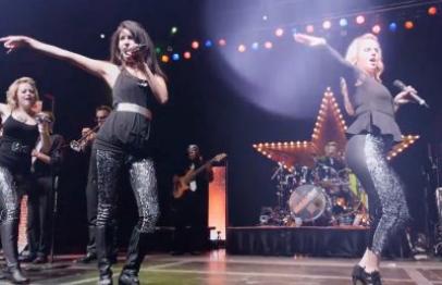 Female singers and dancers perform energetically on stage with a band and colorful lights.