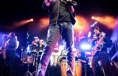 Singer performing energetically on stage with band and colorful lights in the background.