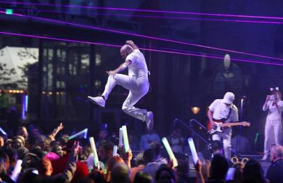 Performer in mid-air jump on stage with colorful lights and enthusiastic crowd below.
