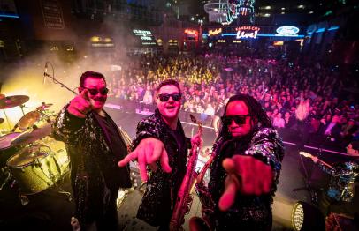 Three musicians in sparkling jackets performing on stage before a cheering crowd.