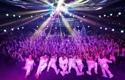 People in white outfits pose on stage, colorful lights shine over a cheering crowd.
