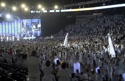 Large indoor event with a crowd dressed in white, bright stage lights, and screens.