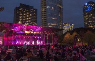 Outdoor concert at night with colorful stage lights and a city skyline in the background.