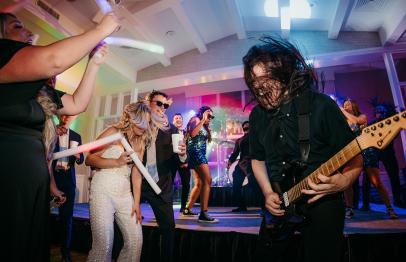 Guitarist performing on stage as people dance and cheer under colorful lights.