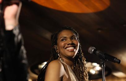 Smiling woman with braids singing into a microphone on stage, warm lighting above.