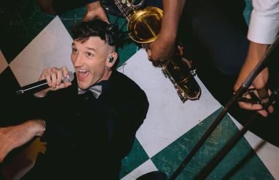 Man in a suit singing on checkered floor with musicians playing brass instruments above him.