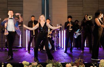 Live band performing on stage with singers and musicians under purple lighting.