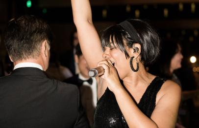 Woman in black dress singing passionately into a microphone at a lively event.