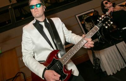 Man in a white tuxedo jacket playing electric guitar on stage.