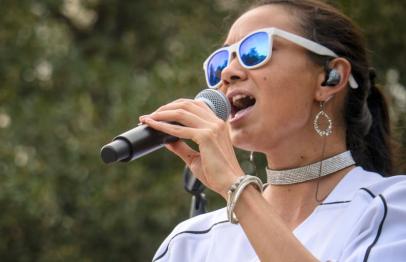Woman wearing sunglasses sings into a microphone outdoors.